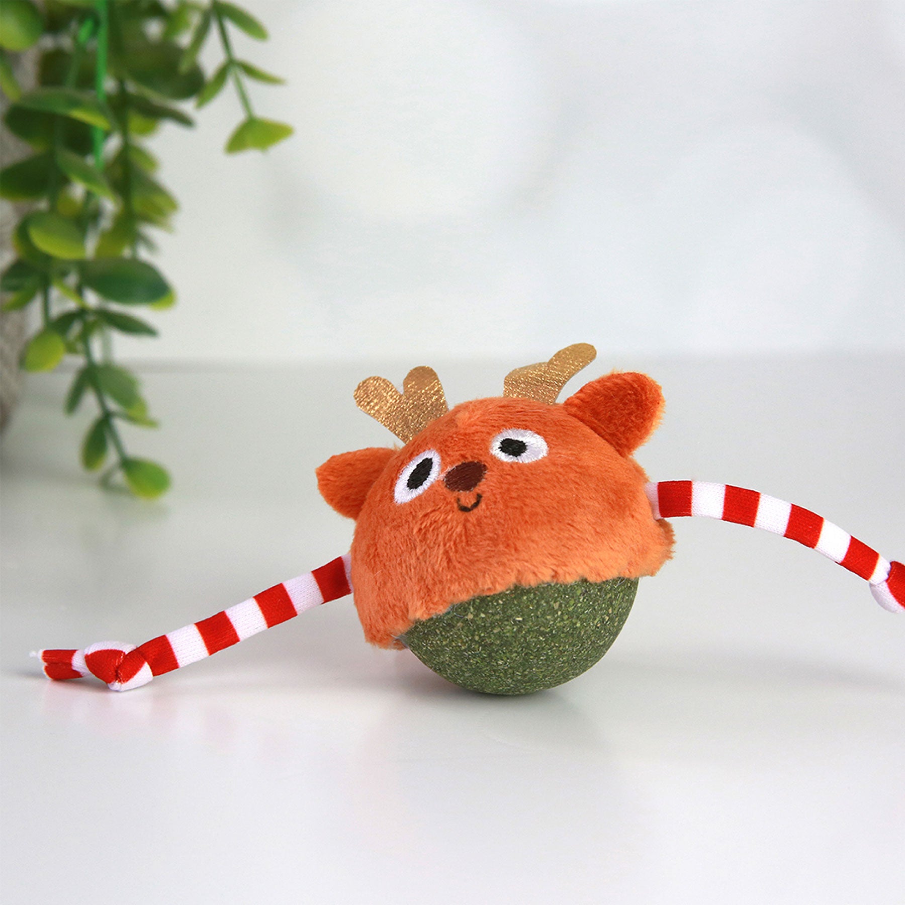 The Mad Cat Catnip Bobbler Roll Around Reindeer toy by Mad Cat has an orange head with antlers and eyes on a green textured catnip stuffed ball, plus red and white striped rope limbs for hunting fun. It rests on a white surface surrounded by green plant leaves.