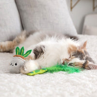 A fluffy cat dozes on a white bed with its head on a pillow. Beside it lies a green feather toy and the Quirky Kitty House Plant Catnip Plush Toy with silvervine leaves, enhancing the rooms cozy vibe.