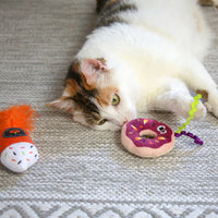 A playful white and orange cat enjoys a Mad Cat Festive Fall Treat Plush Toy on a textured gray carpet with Silvernip sprinkles. Nearby, an orange and white fish-shaped toy awaits, while the plush donut smiles back with purple icing and cheerful sprinkles.