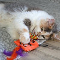 A fluffy cat sprawls on a gray textured rug, playfully biting a Mad Cat Treat Bag Hide-A-Toy, featuring an orange fish design and a purple feather. Infused with catnip, the toy keeps the cats white, gray, and tan fur in constant motion.