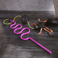 A Mad Cat Creepy Crawly Wand lies on dark wood, featuring a purple curly straw with a decorative black spider with googly eyes and orange-black striped legs. A black spiderweb charm invites interactive play, adding a magical Halloween touch.