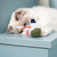 A fluffy white and gray cat, affectionately known as Mad Cat, lounges on a blue surface cuddling a Mad Cat Catnip Bobbler Snowy Day toy. The green mouse wears a blue and white hat with an orange tail, creating a cozy and calm scene.