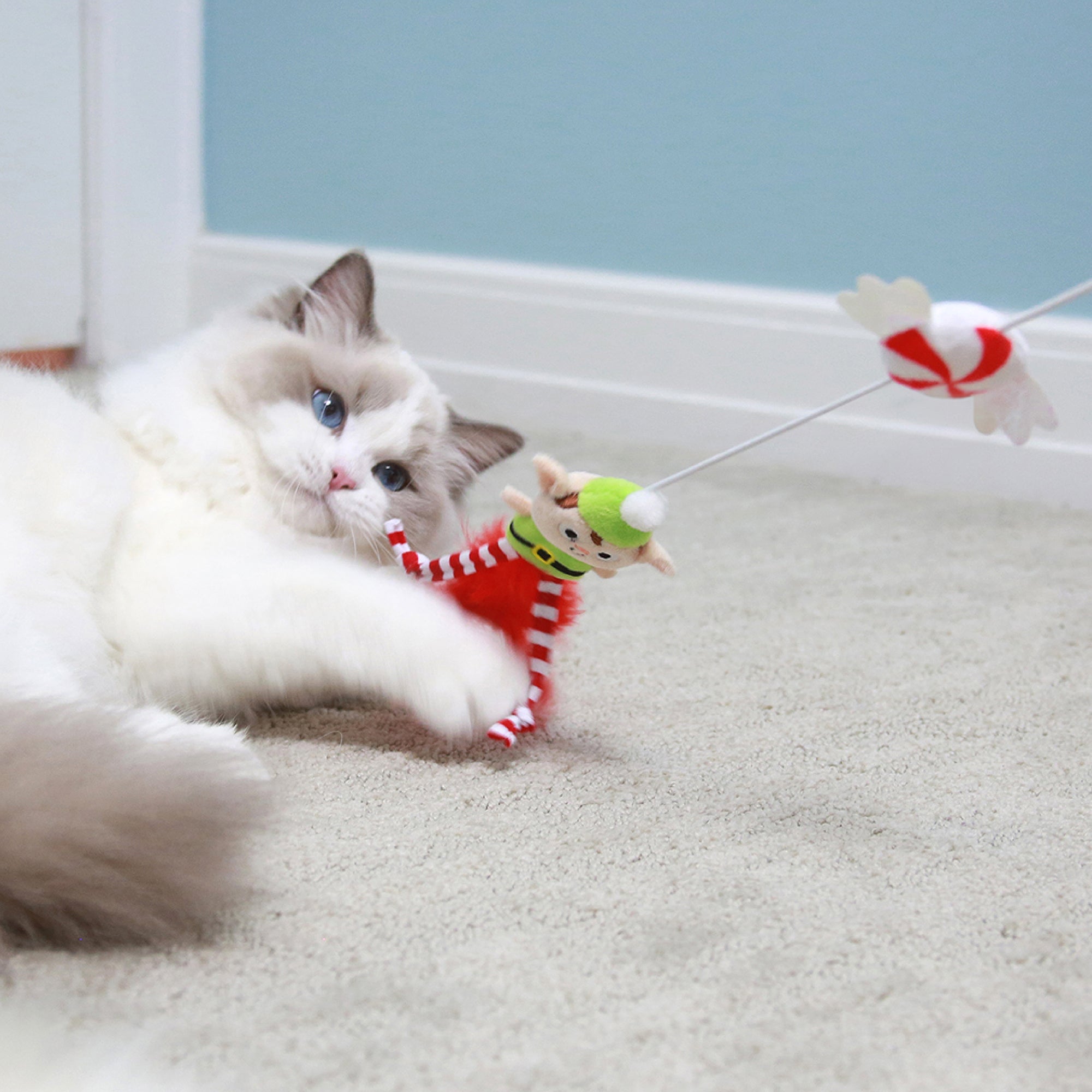 A fluffy cat with blue eyes playfully swings at the Mad Cat Elfin Around Wand Cat Toy, lying relaxed on a light carpet against a blue wall, perfectly capturing the holiday spirit.