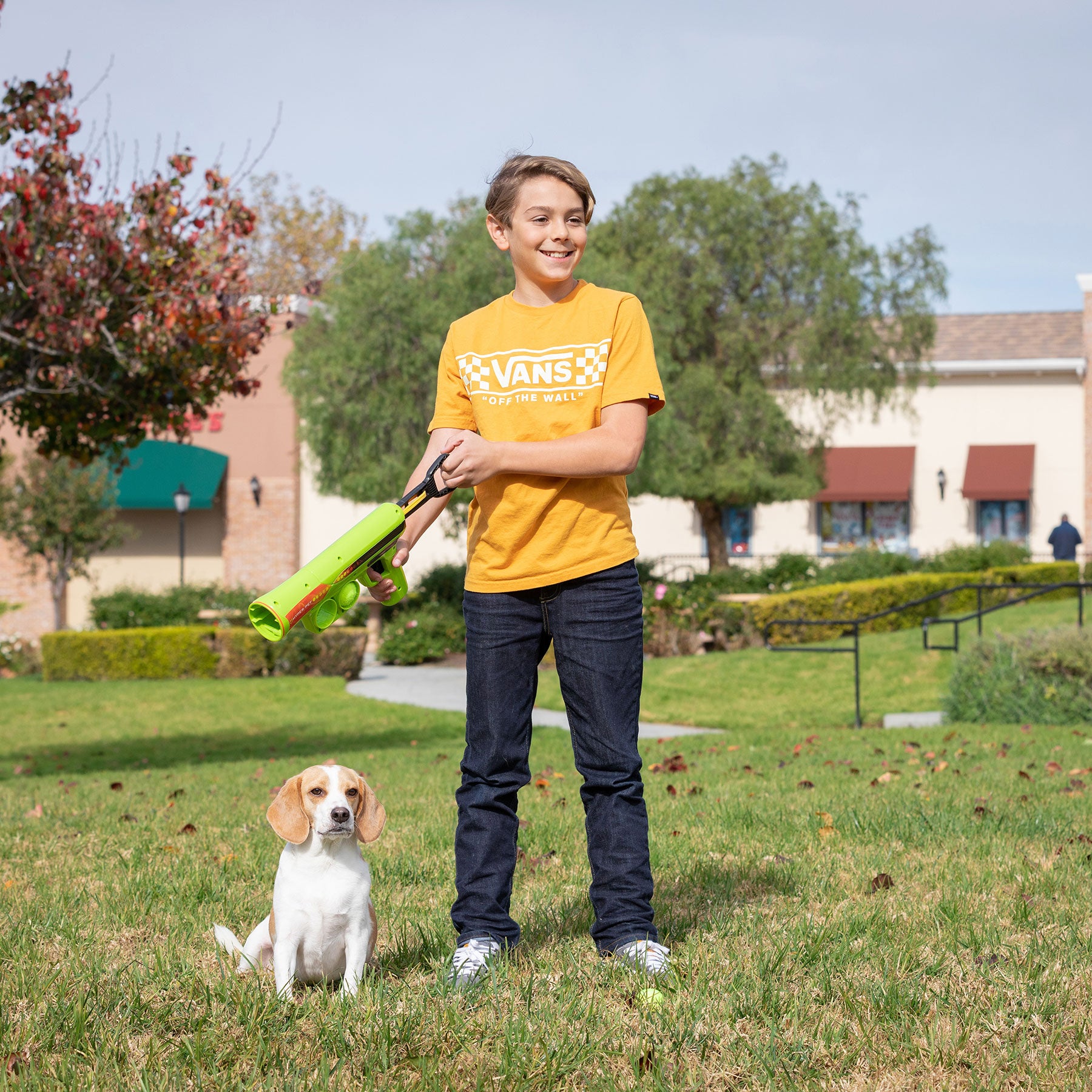 A boy in an orange T-shirt holds a Hyperpet K9K2 Mini Tennis Ball Launcher standing on grass in a park. Beside him sits a beagle, ideal for small dogs. Behind them are trees and buildings with awnings under the cloudy sky.