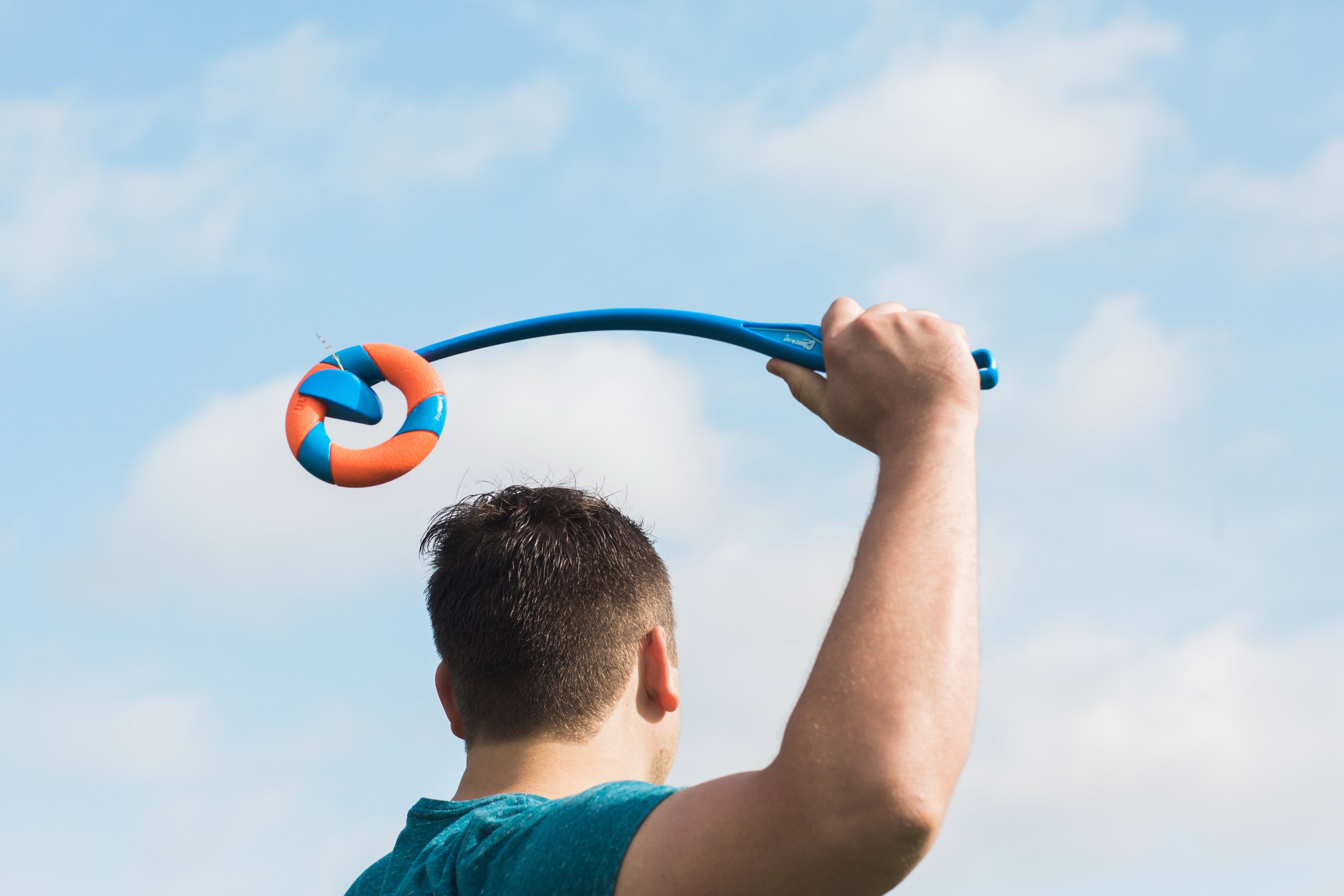 A person in a teal shirt holds the Chuckit! UltraRing Dog Fetch Toy & Launcher Bundle, featuring an orange and blue ball, poised to throw under a clear, partly cloudy sky for an exciting fetch session.