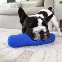 A French bulldog with perked ears and a white and brindle coat playfully chews on a JW Wool-ee Crunch Stick by JW, while lying on a soft, cream-colored rug in the living room.