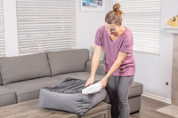 A woman in a purple shirt and gray leggings places a white cushion into a PetFushion Medium Cuddler Bed by Pet Fusion, perfectly matching the cozy living rooms gray sofa, blinds, and picture frame.