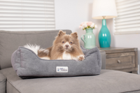 A fluffy brown and white dog rests in a PetFushion Medium Cuddler Bed with high-walled bolsters on a gray couch. A wooden table with a turquoise lamp and flower vase is behind the couch, and window blinds softly light the cozy room.