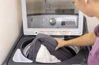 A person in a purple shirt is placing a PetFusion Medium Cuddler Bed, featuring a memory foam base and high-walled bolsters, into an open top-loading washing machine with visible control settings and knobs.